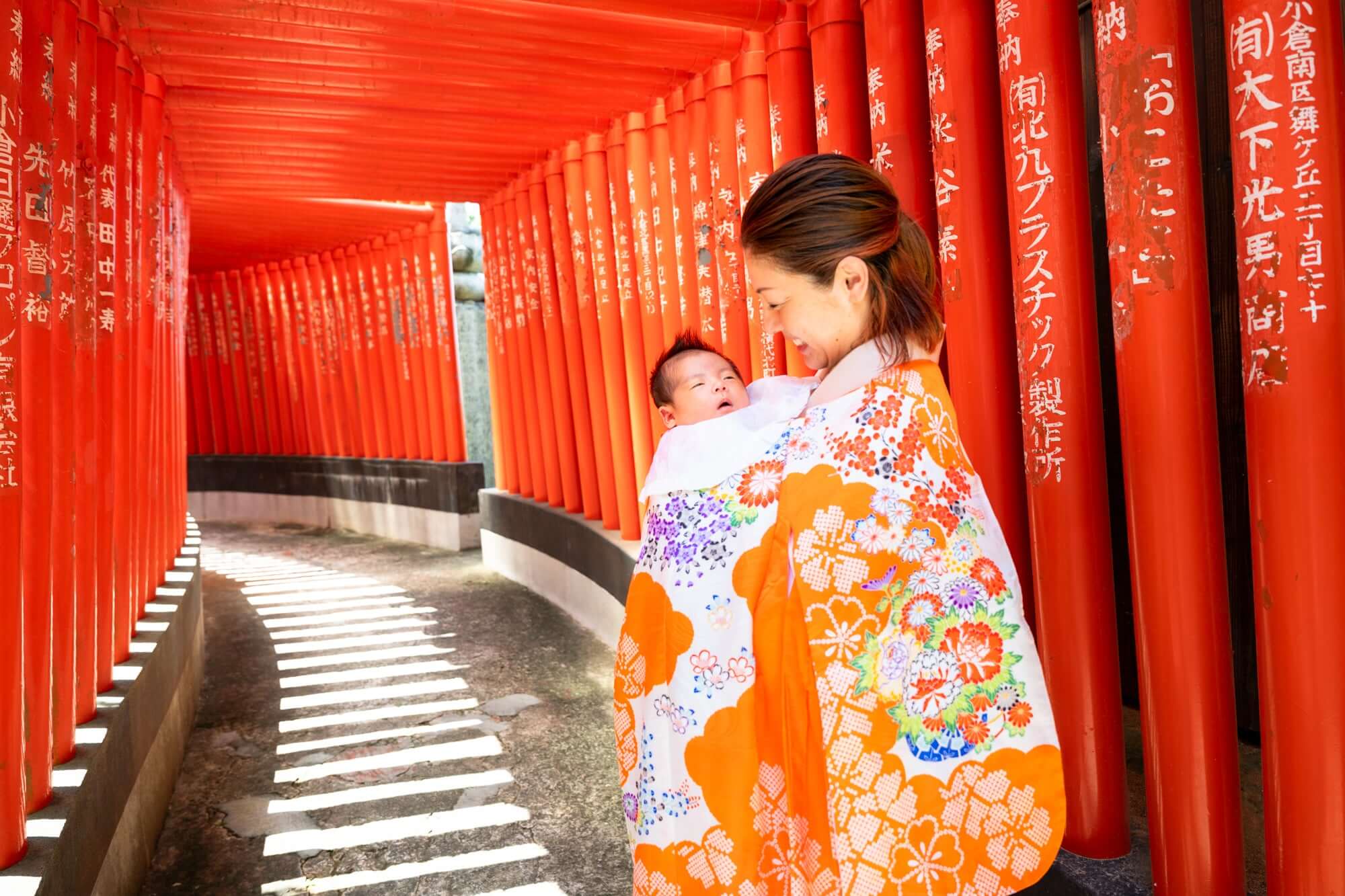 お宮参りの赤ちゃんを抱く母親、赤い鳥居背景にて、フォトウエディング北九州の魅力を表現