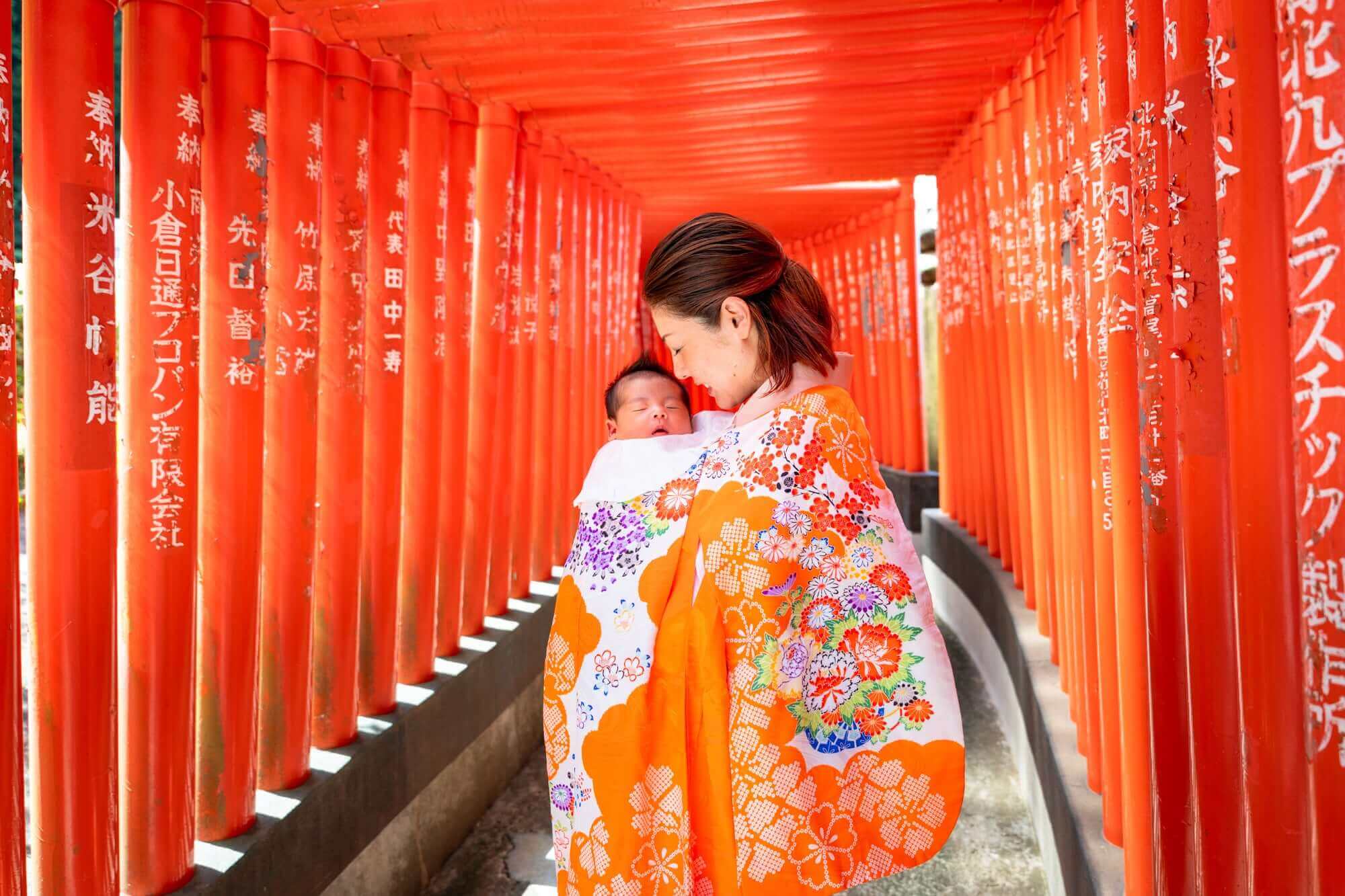 お宮参りの赤ちゃんを抱く母親、鮮やかなオレンジの着物を着用、赤い鳥居の下で撮影。フォトウエディング北九州。